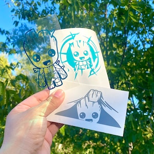 May include: A collection of vinyl stickers featuring the character Groot from Guardians of the Galaxy. The stickers are in various designs and colors, including teal and gray. The stickers are held up against a background of green foliage.