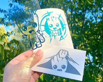 May include: A collection of vinyl stickers featuring the character Groot from Guardians of the Galaxy. The stickers are in various designs and colors, including teal and gray. The stickers are held up against a background of green foliage.