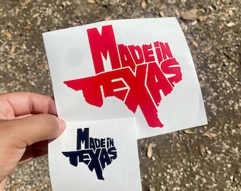 Puede incluir: Dos pegatinas blancas con el texto "Made in Texas" en una fuente estilizada. La pegatina más grande es roja y la más pequeña es azul marino. El texto tiene la forma del estado de Texas.