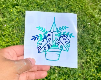May include: A blue and green vinyl sticker featuring a hanging potted plant design. The plant has large, stylized leaves and is suspended by a thin line. The sticker is held against a background of green grass.
