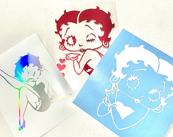 Puede incluir: Tres pegatinas de vinilo con Betty Boop en varias poses. Una pegatina es holográfica, otra es roja y la tercera es blanca sobre un fondo azul. Las pegatinas están troqueladas y listas para aplicar.