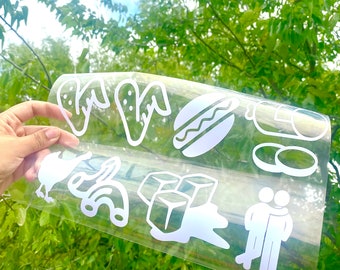 Puede incluir: Una lámina transparente con recortes de vinilo blanco de iconos de comida y sociales. Los iconos incluyen alitas de pollo, un perrito caliente, una botella, dos rodajas de pepinillos, un pavo, macarrones, cubitos de hielo y dos figuras abrazándose.