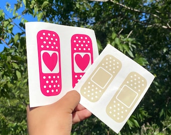 May include: Two sheets of stickers are held up against a background of green leaves and blue sky. One sheet has two pink bandages with a heart in the center. The other sheet has two beige bandages.