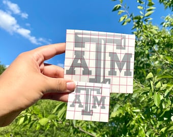 Puede incluir: Dos calcomanías de vinilo grises con las letras "ATM" en una fuente universitaria. Las calcomanías están sobre un respaldo de cuadrícula, una más grande y otra más pequeña. El fondo es un cielo azul brillante y follaje verde.