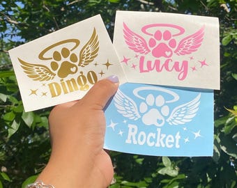 May include: Three pet memorial decals with paw prints, wings, and halos. One is gold with "Dingo", another is pink with "Lucy", and the last is blue with "Rocket". Each has star accents.