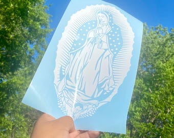 May include: A light blue vinyl decal featuring a white illustration of the Virgin Mary. The image depicts Mary with her hands clasped in prayer, surrounded by a halo of stars and rays. The decal is held against a backdrop of a bright blue sky and green trees.