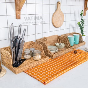 May include: Three woven baskets with a checkered orange and white runner on a white countertop. The baskets are filled with various items, including kitchen knives, jars, and dishes. The text "VATIMA HANDMADE WITH LOVE" is visible on the wall behind the baskets.