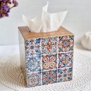 May include: A square tissue box with a wooden top and sides adorned with colourful, patterned tiles. White tissues protrude from the top opening. The tiles display blue, orange, and yellow floral patterns.