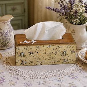 Puede incluir: Una caja de pañuelos rectangular con tapa de madera oscura que presenta un motivo de pájaro y flores blancas. Los laterales están decorados con un patrón vintage beige de pequeñas flores azules, amarillas y verdes sobre un fondo texturizado.