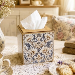 Puede incluir: Una caja de pañuelos cuadrada con un detallado patrón floral de damasco en tonos azul, marrón y crema. La caja tiene una tapa de madera plana con una abertura rectangular central, de la cual sobresale un pañuelo blanco hacia arriba.
