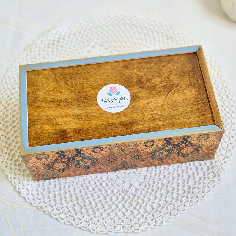 Pu&ograve; includere: Una scatola di legno con un bordo blu chiaro e dorato. La parte superiore della scatola &egrave; tinta di un colore marrone scuro. Un adesivo circolare bianco con un fiore rosa e il testo "BARVY gifts" &egrave; sulla parte superiore della scatola. La parte inferiore della scatola ha un design a motivi.