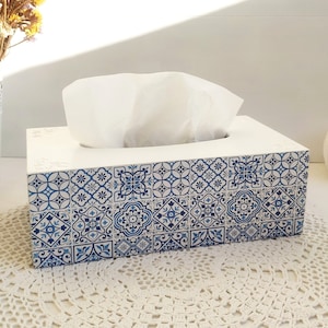 Puede incluir: Caja de pañuelos rectangular blanca con un patrón de azulejos azules y blancos. La caja tiene un diseño floral decorativo en la parte superior y está llena de pañuelos blancos. La caja de pañuelos está sobre un tapete de ganchillo blanco.