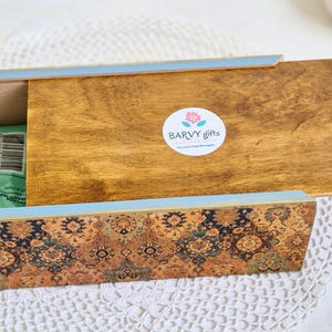 Pu&ograve; includere: Un porta fazzoletti in legno con un bordo blu e un design a motivi. Il coperchio &egrave; aperto e l'interno &egrave; rivestito con un tessuto blu chiaro. La scatola ha un adesivo sulla parte superiore che dice "Barvy Gifts".