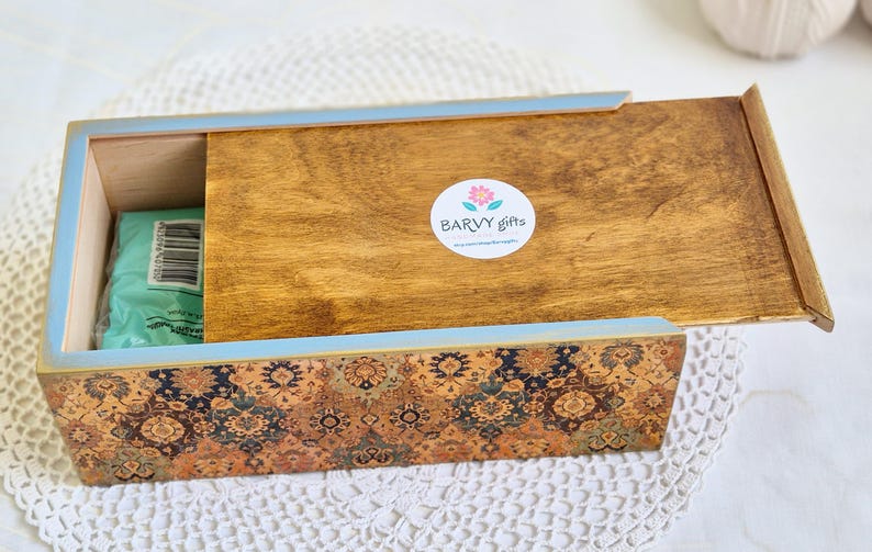 Pu&ograve; includere: Un porta fazzoletti in legno con un bordo blu e un design a motivi. Il coperchio &egrave; aperto e l'interno &egrave; rivestito con un tessuto blu chiaro. La scatola ha un adesivo sulla parte superiore che dice "Barvy Gifts".