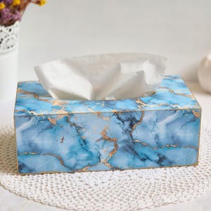 Puede incluir: Una caja de pañuelos rectangular con un patrón de mármol azul y blanco con vetas doradas metálicas. La caja tiene una abertura rectangular en la parte superior de la que sobresale un pañuelo blanco, con un acabado liso.