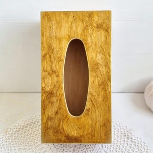 Pu&ograve; includere: Un porta fazzoletti in legno con una finitura marrone dorata. La scatola ha una forma rettangolare con un'apertura ovale arrotondata per i fazzoletti.