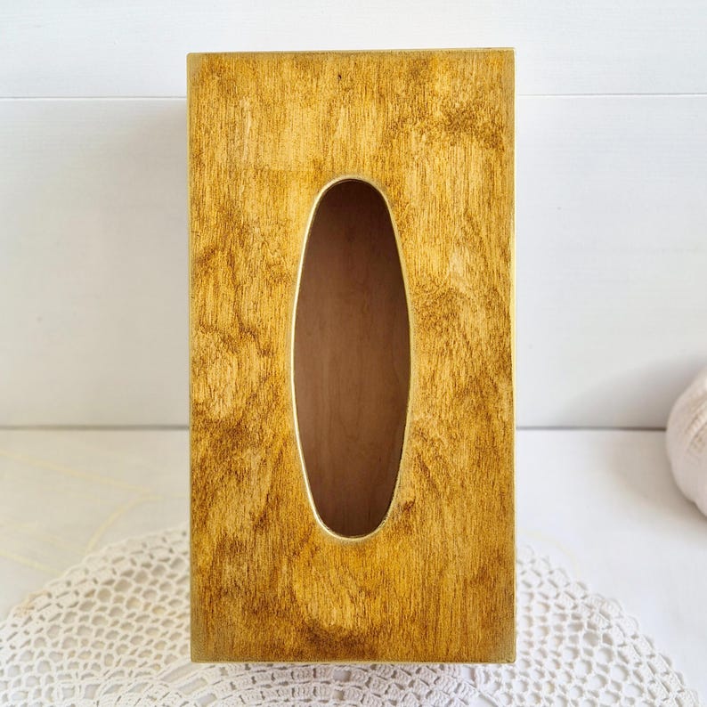 Pu&ograve; includere: Un porta fazzoletti in legno con una finitura marrone dorata. La scatola ha una forma rettangolare con un'apertura ovale arrotondata per i fazzoletti.