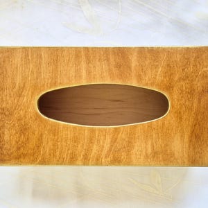 Op de afbeelding: Een rechthoekige houten tissuebox met een natuurlijke houtnerf afwerking. De doos heeft een ovale opening in het midden voor het afgeven van tissues. Het hout heeft een warme, goudbruine tint.