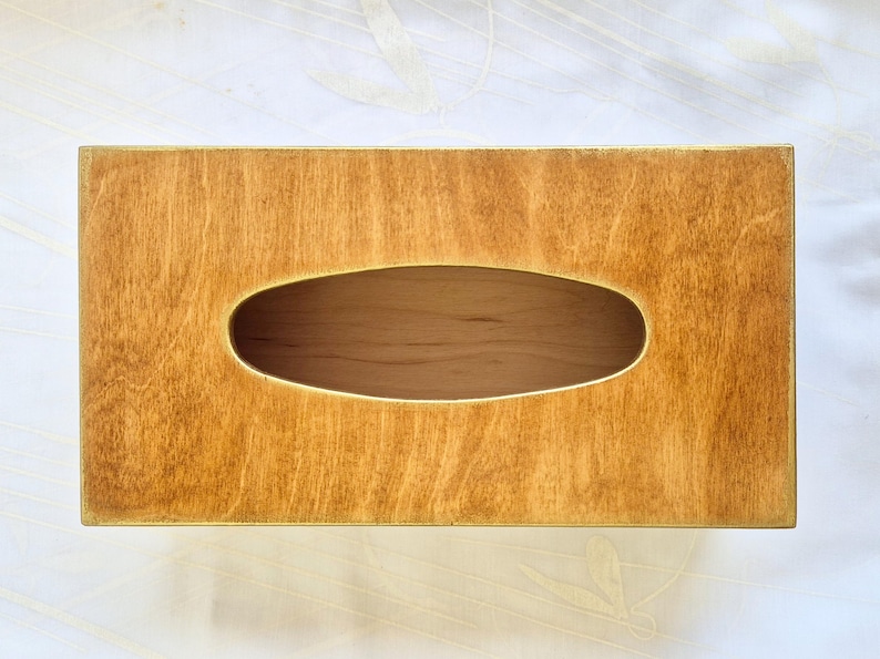 Op de afbeelding: Een rechthoekige houten tissuebox met een natuurlijke houtnerf afwerking. De doos heeft een ovale opening in het midden voor het afgeven van tissues. Het hout heeft een warme, goudbruine tint.