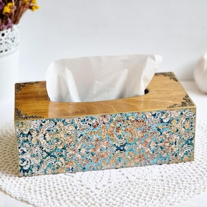 Pode incluir: Um porta-lenços de madeira com um design decorativo de padrão azul, dourado e branco. A tampa tem uma ranhura para lenços e uma borda dourada.