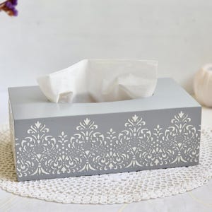 May include: A grey rectangular tissue box with a white, decorative floral pattern along the bottom. White tissues protrude from the top. The box rests on a white, crocheted doily.