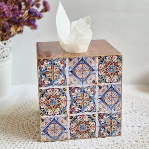 May include: A wooden tissue box with a decorative tile design. The box has a light brown wooden top and sides decorated with colourful, patterned tiles in blue, red, yellow, and white. White tissues are visible at the top.