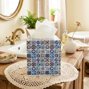 May include: A square tissue box cover featuring a repeating pattern of blue, white, and gold geometric tiles. The box is topped with white tissues and sits on a round, cream-coloured crocheted doily on a wooden surface.