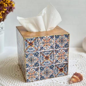 May include: A square tissue box with a wooden lid and sides decorated with patterned tiles. The tiles display a repeating design in blue, orange, and white. White tissues protrude from the top. The box rests on a white lace doily.