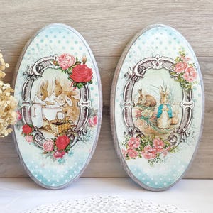 Può includere: Due targhe da parete ovali in legno con illustrazioni decorative di conigli. La targa sinistra mostra conigli con una brocca e un cestino, mentre quella destra raffigura un coniglio con una giacca blu. Entrambe hanno bordi decorati e rose.