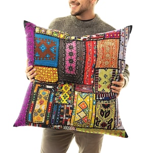 May include: A large, square patchwork cushion with a vibrant mix of colours and patterns. The cushion features a variety of fabric pieces in shades of purple, blue, yellow, and red, creating a unique and textured design. The cushion is held up by a person.