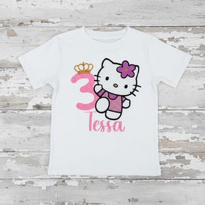 May include: White t-shirt featuring a pink number 3 with a gold crown, a cartoon cat character with a pink flower, and the name "Tessa" in pink script. The cat is wearing a pink dress.