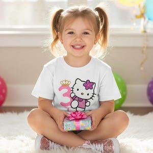 May include: A white t-shirt featuring a graphic of Hello Kitty with a pink flower and the number 3 with a crown. The shirt is worn by a young child holding a wrapped gift. The child is sitting on a white rug.
