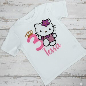 May include: White t-shirt featuring a glittery pink number 3 with a gold crown, a cartoon cat character, and the name "Tessa" in pink glitter. The cat has a purple flower and is wearing a purple dress.