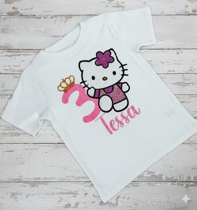 May include: White t-shirt featuring a glittery pink number 3 with a gold crown, a cartoon cat character, and the name "Tessa" in pink glitter. The cat has a purple flower and is wearing a purple dress.