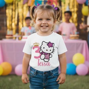 May include: A young girl wearing a white t-shirt with a graphic of Hello Kitty and the number 3. The shirt also has the name "Tessa" printed on it. She is wearing blue jeans and has purple hair bows. The background includes party decorations.