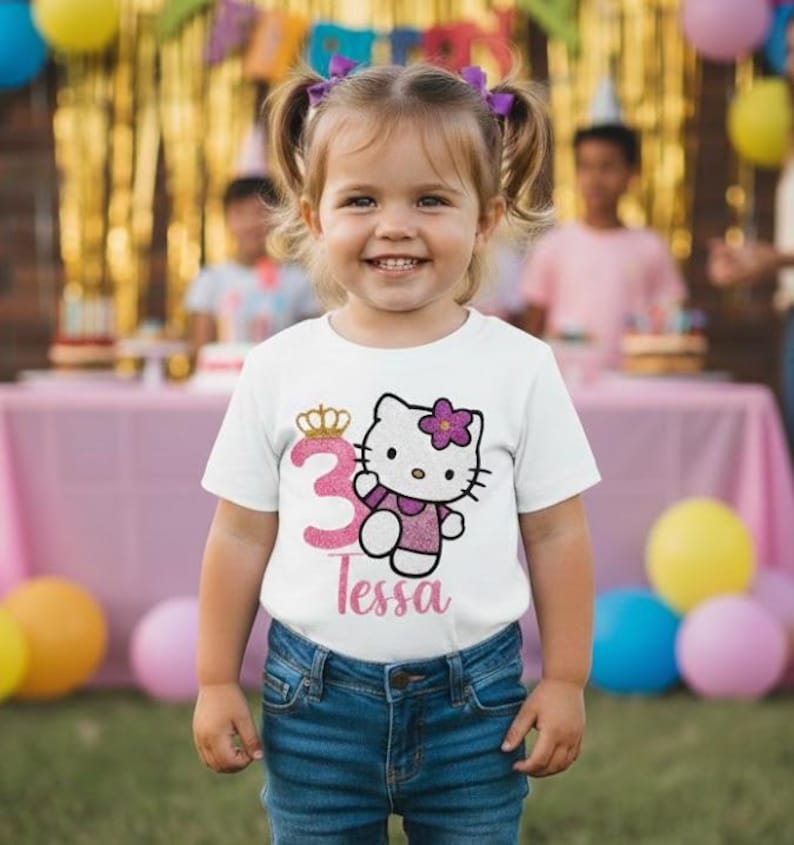 May include: A young girl wearing a white t-shirt with a graphic of Hello Kitty and the number 3. The shirt also has the name "Tessa" printed on it. She is wearing blue jeans and has purple hair bows. The background includes party decorations.