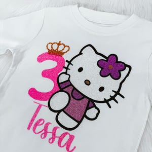 May include: White t-shirt featuring a glittery pink number 3 with a gold crown, a Hello Kitty design with a purple flower, and the name "Tessa" in pink glitter. The shirt is designed for a child's birthday.