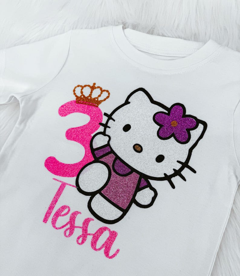 May include: White t-shirt featuring a glittery pink number 3 with a gold crown, a Hello Kitty design with a purple flower, and the name "Tessa" in pink glitter. The shirt is designed for a child's birthday.