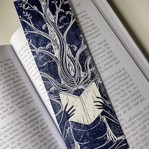 May include: A blue and white linocut print of a person reading a book. The person is sitting with their legs crossed and their head is surrounded by a tree with leaves. The print is on a white background.