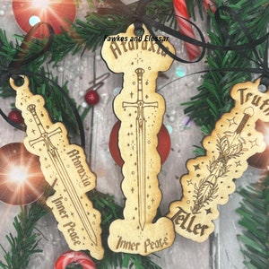 May include: Three wooden Christmas decorations featuring a sword design. The ornaments are decorated with stars and the words "Ataraxia Inner Peace", "Truth Teller Inner Peace", and "Fawkes and Elessar Ataraxia Inner Peace".