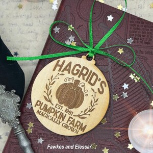 May include: A round wooden bauble with a brown pumpkin and green leaves. The bauble says "Hagrid's Pumpkin Farm Est. 1981 Magically Grown" in a brown font.
