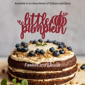 May include: A red glitter cake topper with the words "little pumpkin" and a small pumpkin design. The topper is on a cake with blueberries and walnuts.