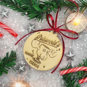 May include: A circular wooden Christmas ornament featuring the text "Griswold's Famous Eggnog" and a cartoon moose design. The ornament is tied with a red ribbon and surrounded by festive decorations, including candy canes and silver stars.