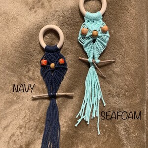 Puede incluir: Dos colgantes de pared de búho de macramé con anillos de madera. El búho de la izquierda es azul marino con cuentas naranjas y marrones para los ojos. El búho de la derecha es azul claro con cuentas marrones para los ojos. El texto "NAVY" está debajo del búho de la izquierda y el texto "SEAFOM" está debajo del búho de la derecha.