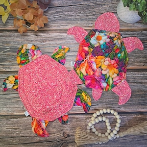 May include: Two turtle-shaped fabric items. One features a vibrant floral pattern with stained-glass style outlines, while the other is covered in a bright pink glittery fabric. Both have four flippers and a head, with a small white tag on the side.