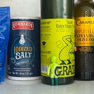 May include: A blue Lombardi Iodized Salt container, a Grazia olive oil bottle with a bright yellow label, a Carapelli olive oil bottle, and a small, neon green figurine with a black letter G standing in front of the Grazia bottle.