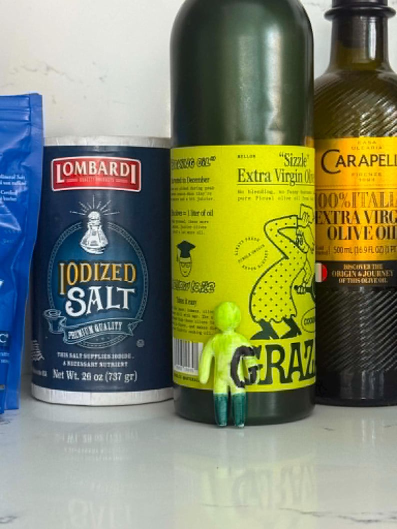 May include: A blue Lombardi Iodized Salt container, a Grazia olive oil bottle with a bright yellow label, a Carapelli olive oil bottle, and a small, neon green figurine with a black letter G standing in front of the Grazia bottle.