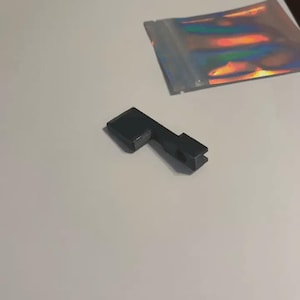 May include: A small, matte black, L-shaped component with a rectangular base and a narrow, notched arm featuring a circular opening. Beside it lies a small, rectangular, iridescent holographic pouch with a metallic finish and a zip-lock seal.