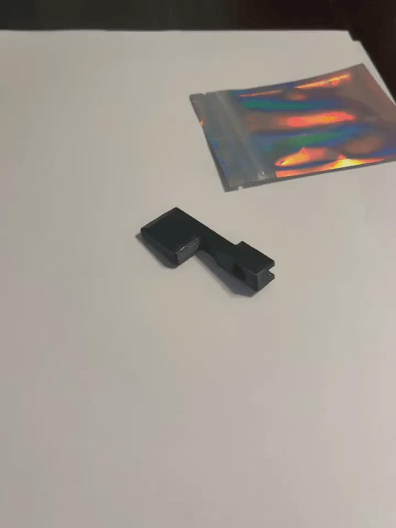 May include: A small, matte black, L-shaped component with a rectangular base and a narrow, notched arm featuring a circular opening. Beside it lies a small, rectangular, iridescent holographic pouch with a metallic finish and a zip-lock seal.