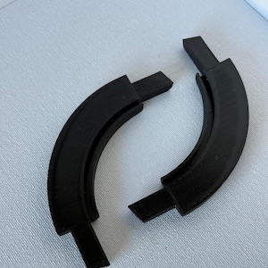 May include: Two black, curved, 3D-printed plastic components with rectangular tabs extending from each end. The pieces feature a textured, layered surface finish consistent with additive manufacturing processes and have a hollowed-out inner channel.